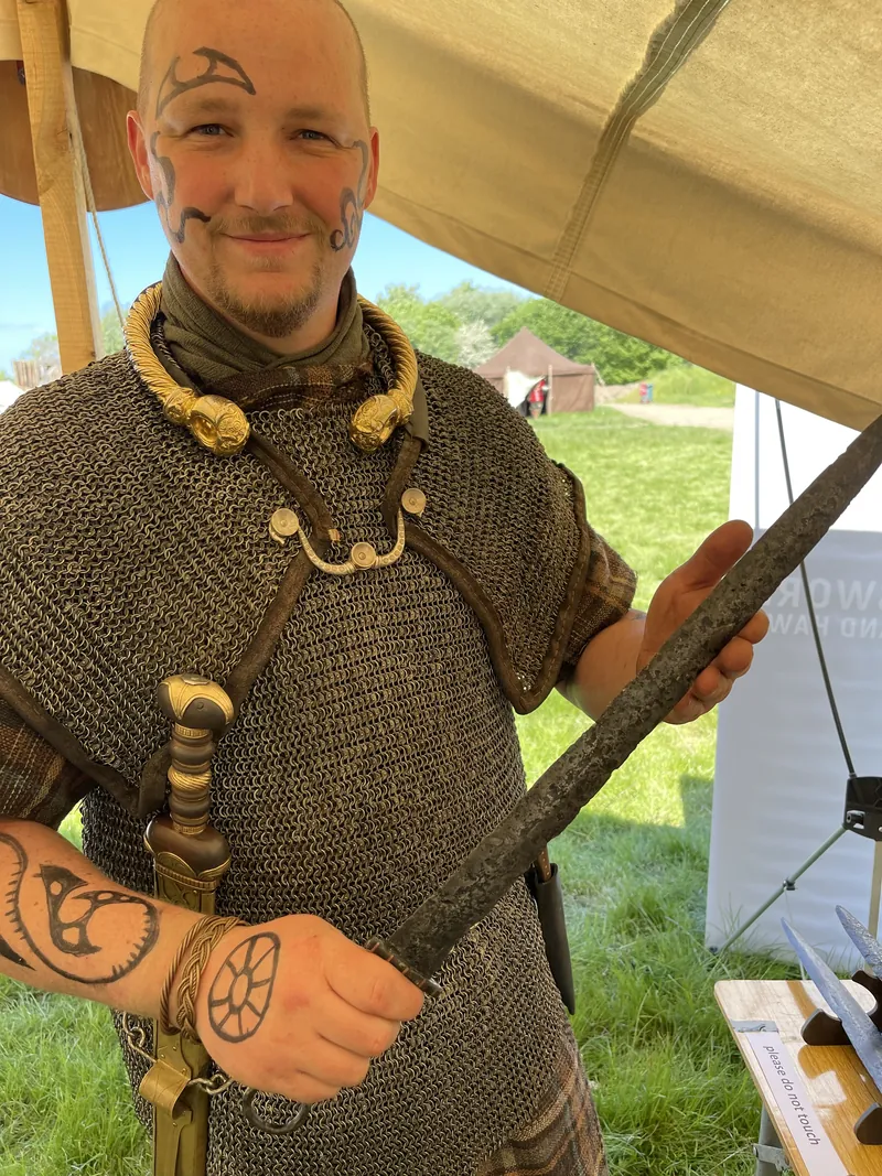 A re-enactor in a mail shirt and heavy neck-torc holding a spear beneath an awning