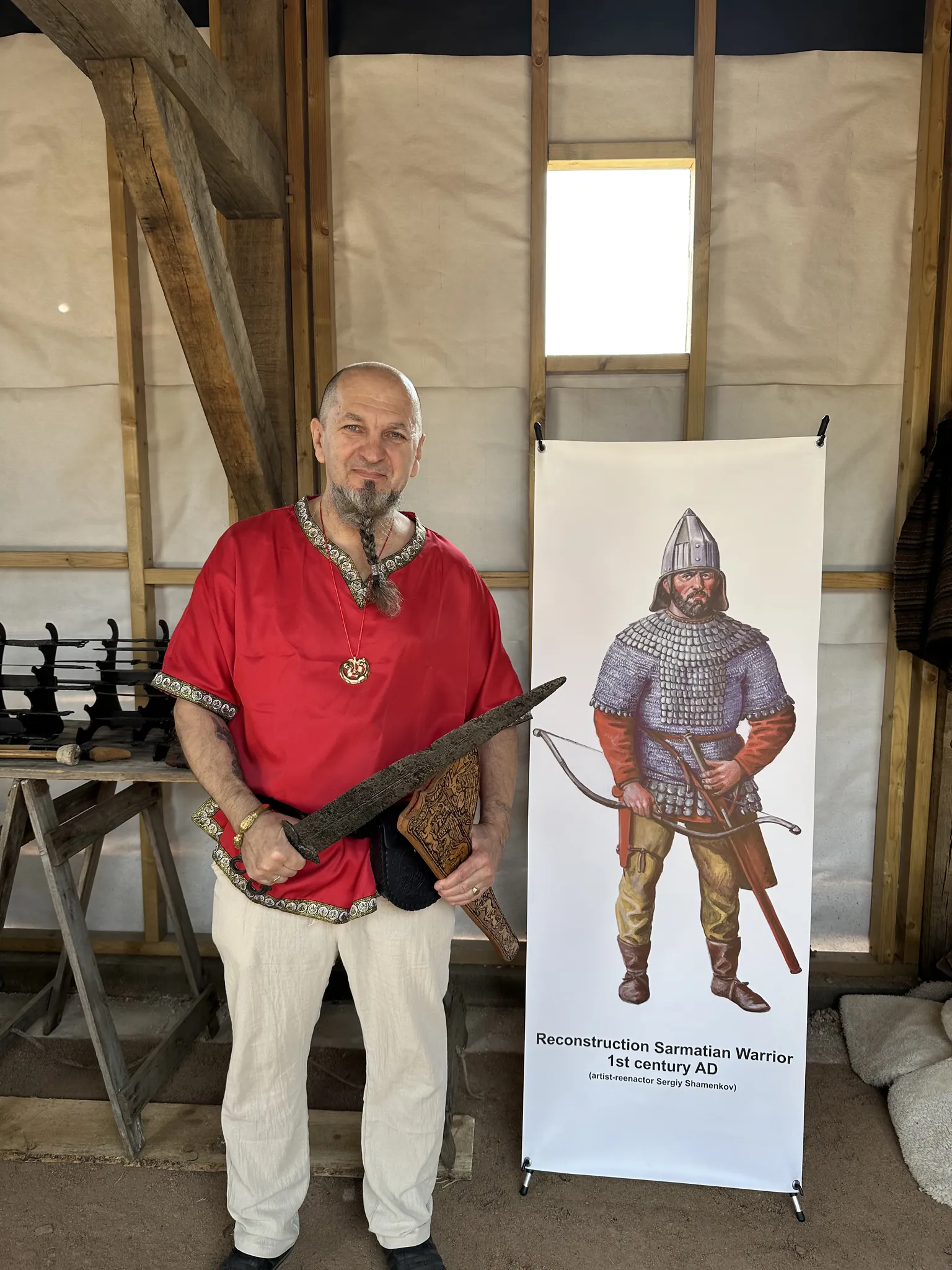 A Society member in a red tunic holding a sheathed sword beside the Sarmatian banner