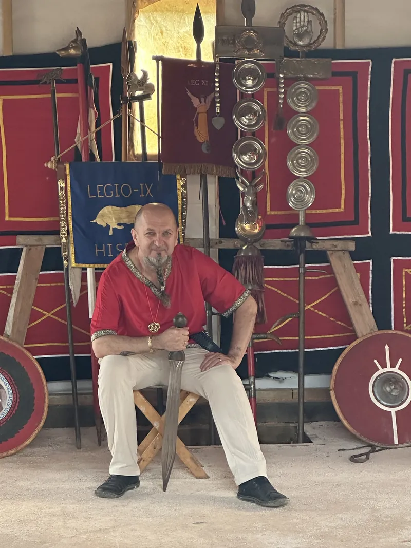 A man seated before the hanging banners and painted shields of Legio IX
