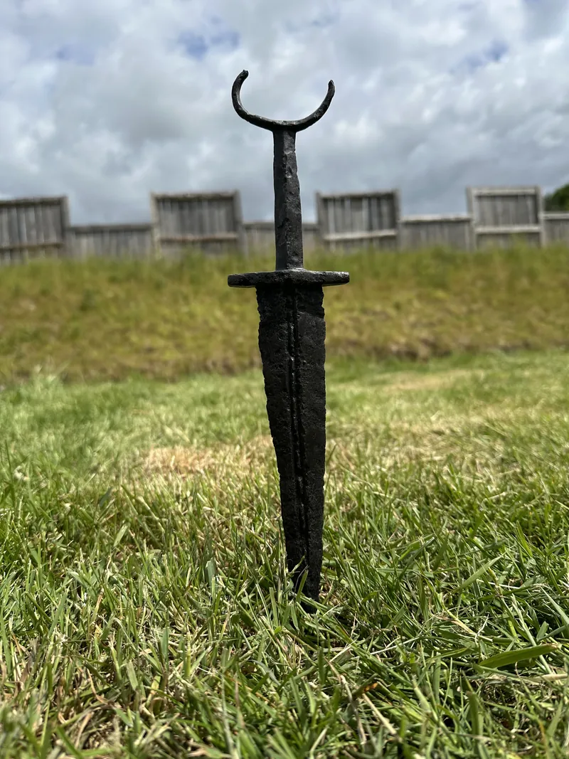 An iron sword standing point-down in the grass before the fort palisade
