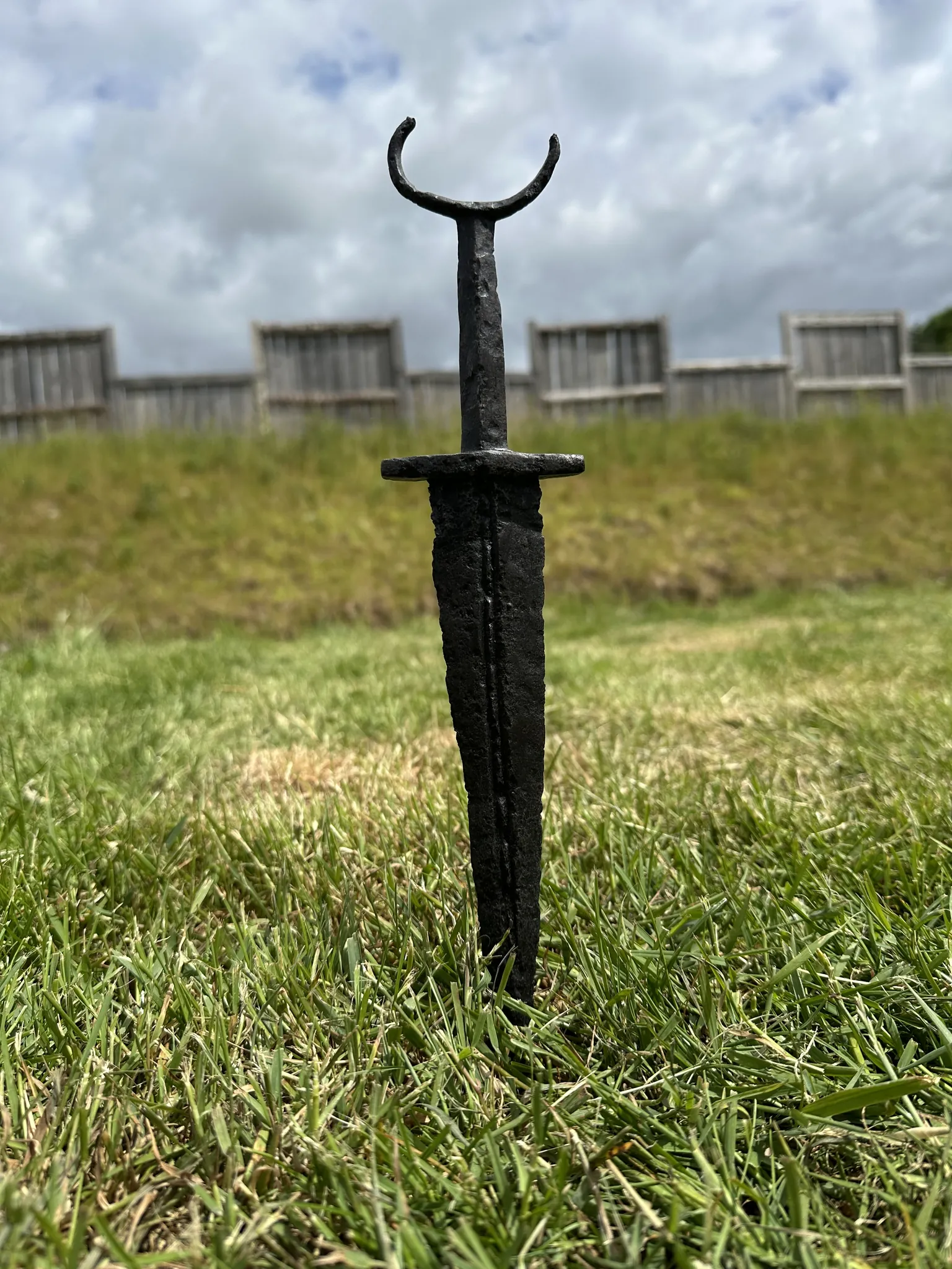 An iron sword standing point-down in the grass before the fort palisade