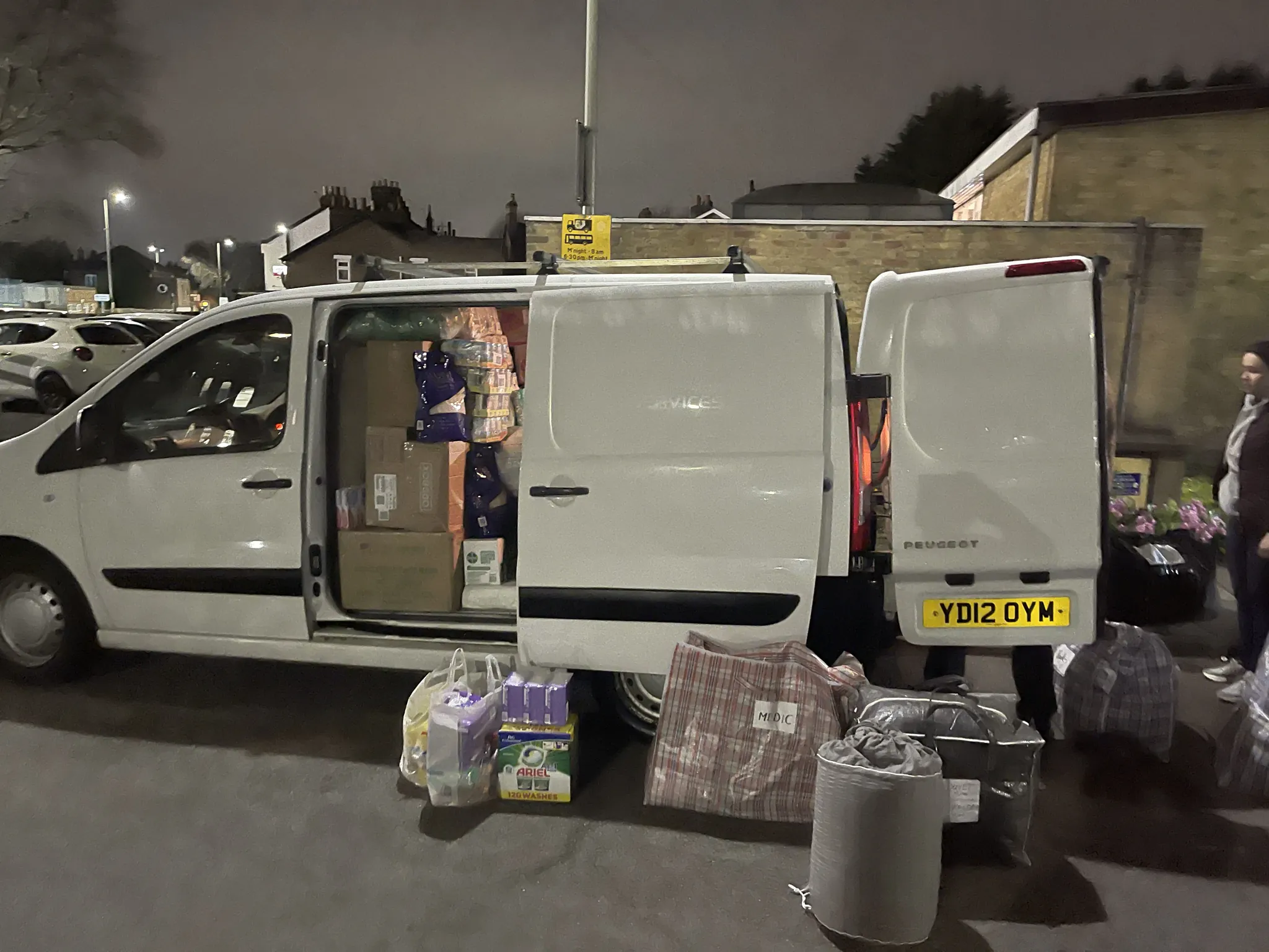 A white van with its rear doors open at dusk, aid bags and bottled water stacked on the tarmac beside it