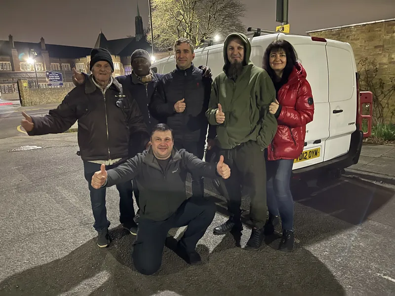 Six volunteers in winter coats give a thumbs-up beside a loaded van after dark